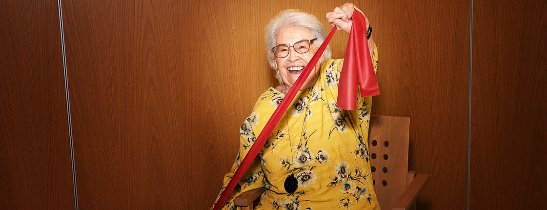 ältere Frau macht Gymnastik Übungen mit einem Tuch. Sie sitzt auf einem Stuhl vor einer braunen Wand