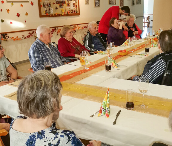 Eine Gruppe von Menschen, die an einem festlich gedeckten Tisch sitzen. Die Tischdekoration ist herbstlich gestaltet, passend zum Erntedankfest. Die Personen wirken entspannt und gesellig, während sie gemeinsam Zeit verbringen.