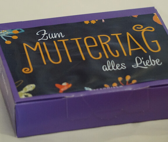 Eine lila Schachtel mit Blumenmuster mit der Aufschrift "Zum Muttertag alles Liebe"
