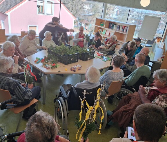 Bewohnerinnen und Bewohner des Pflegewohnhauses sitzen im großen Gemeinschaftsraum zusammen und binden Palmbuschen. In der Mitte des Raumes stehen Kisten voller frischer Zweige, während die Gruppe in geselliger Runde bastelt, plaudert und sich auf das bevorstehende Palmwochenende vorbereitet.