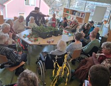 Bewohnerinnen und Bewohner des Pflegewohnhauses sitzen im großen Gemeinschaftsraum zusammen und binden Palmbuschen. In der Mitte des Raumes stehen Kisten voller frischer Zweige, während die Gruppe in geselliger Runde bastelt, plaudert und sich auf das bevorstehende Palmwochenende vorbereitet.