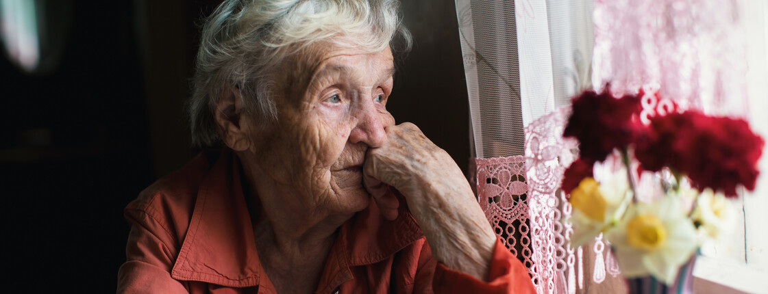 Portrait einer Seniorin, die an einem kleinen Küchentisch sitzt, ihren Kopf auf dem linken Arm abstützt und aus dem Fenster schaut. Auf dem Tisch stehen Blumen in einer Vase.