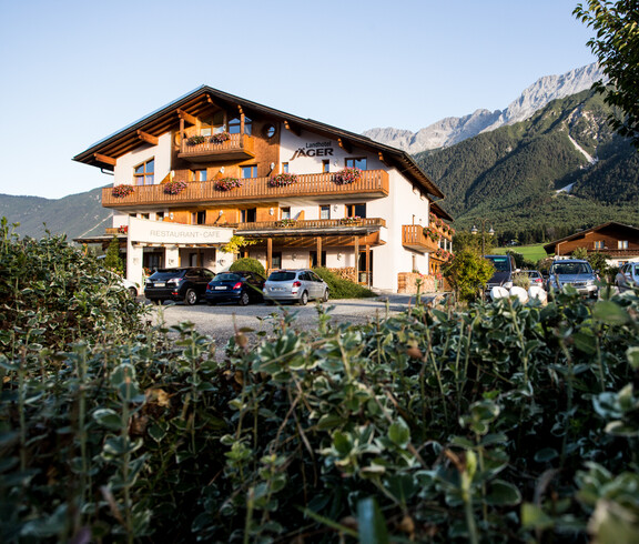 Ein traditionelles Hotel in den Alpen mit einem asphaltierten Parkplatz. Im Vordergrund befindet sich eine grüne Hecke.