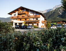 Ein traditionelles Hotel in den Alpen mit einem asphaltierten Parkplatz. Im Vordergrund befindet sich eine grüne Hecke.