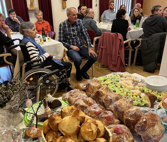 Bewohnerinnen und Bewohner sowie Gäste sitzen im Gemeinschaftsraum eines Pflegewohnhauses, im Vordergrund steht ein Kuchen‑ und Gebäckbuffet