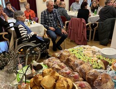 Bewohnerinnen und Bewohner sowie Gäste sitzen im Gemeinschaftsraum eines Pflegewohnhauses, im Vordergrund steht ein Kuchen‑ und Gebäckbuffet