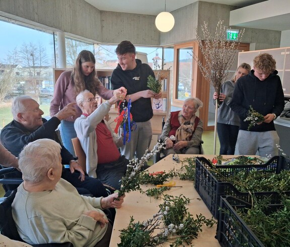 Im Gemeinschaftsraum des Pflegewohnhauses arbeiten Bewohnerinnen und Bewohner gemeinsam mit jungen Helferinnen und Helfern an bunten Palmbuschen. Zweige und Materialien liegen auf den Tischen bereit.