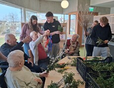 Im Gemeinschaftsraum des Pflegewohnhauses arbeiten Bewohnerinnen und Bewohner gemeinsam mit jungen Helferinnen und Helfern an bunten Palmbuschen. Zweige und Materialien liegen auf den Tischen bereit.