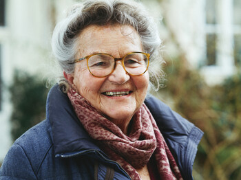 Portrait einer Seniorin mit Brille, die einen roten Schal und einen blauen Wintermantel trägt.