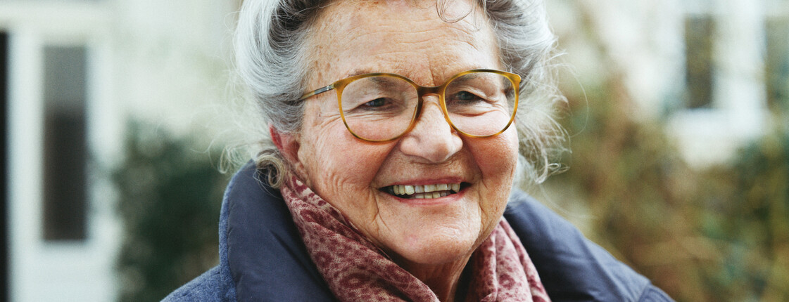 Fachstelle für Pflegende Angehörige Portrait einer Seniorin mit Brille, die einen roten Schal und einen blauen Wintermantel trägt.