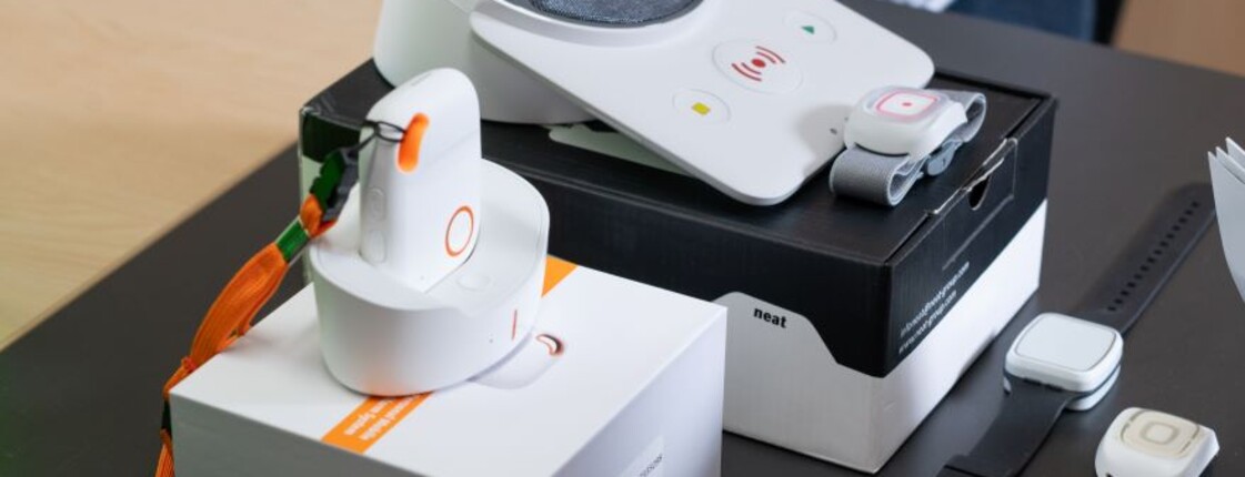 Mehrere Komponenten des Caritas-Notrufsystems liegen auf einem Tisch: eine Basisstation, mobile Notrufgeräte mit orangefarbenem Knopf in Ladestationen, Armband- und Anhänger-Notrufknöpfe sowie Verpackungen und ein Informationsfolder „Caritas Pflege Notruftelefon“.
