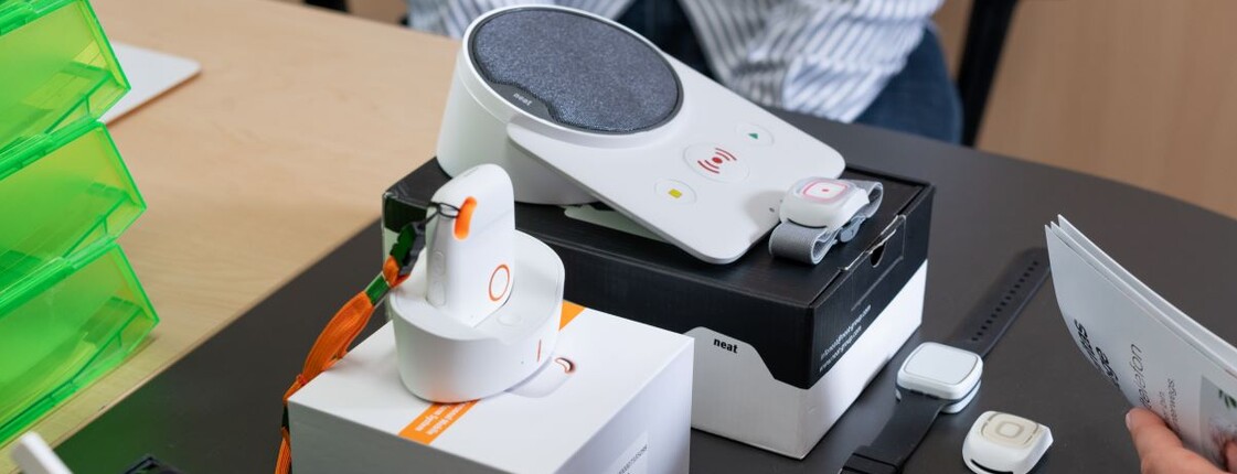 Mehrere Komponenten des Caritas-Notrufsystems liegen auf einem Tisch: eine Basisstation, mobile Notrufgeräte mit orangefarbenem Knopf in Ladestationen, Armband- und Anhänger-Notrufknöpfe sowie Verpackungen und ein Informationsfolder „Caritas Pflege Notruftelefon“.