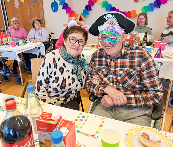 Ein Paar feiert fröhlich bei der Faschingsfeier im Haus Lisa. Der Mann trägt einen Piratenhut und eine bunte Maske, die Frau trägt weiße Katzenohren. Im Hintergrund sitzen weitere Gäste, umgeben von bunten Dekorationen.