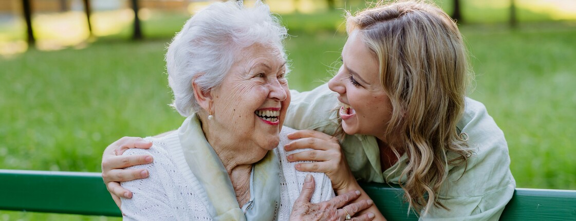 Demenz-Servicezentrum  In einem Park sitzt eine Seniorin auf einer grünen Bank. Hinter ihr steht eine junge Frau, die ihre Hände auf die Schultern der Seniorin gelegt hat. Beide schauen sich lachend an.