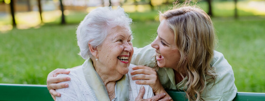 Demenz-Servicezentrum In einem Park sitzt eine Seniorin auf einer grünen Bank. Hinter ihr steht eine junge Frau, die ihre Hände auf die Schultern der Seniorin gelegt hat. Beide schauen sich lachend an.