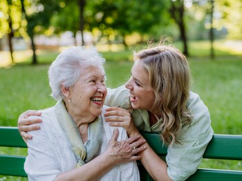 Demenz-Servicezentrum  In einem Park sitzt eine Seniorin auf einer grünen Bank. Hinter ihr steht eine junge Frau, die ihre Hände auf die Schultern der Seniorin gelegt hat. Beide schauen sich lachend an.