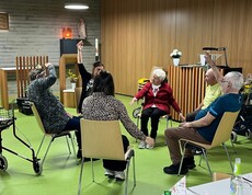 Mehrere Bewohner*innen des Pflegewohnhauses sitzen in einem Stuhlkreis und führen gemeinsam mit einer anleitenden Person Bewegungsübungen mit den Armen durch.