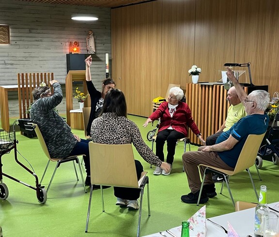 Mehrere Bewohner*innen des Pflegewohnhauses sitzen in einem Stuhlkreis und führen gemeinsam mit einer anleitenden Person Bewegungsübungen mit den Armen durch.
