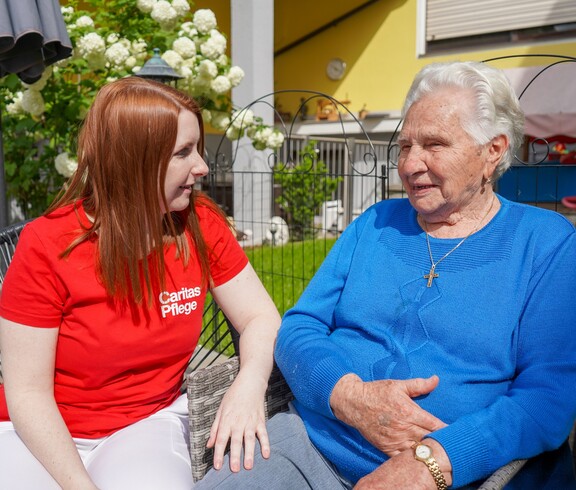 Eine Caritas Pflegerin sitzt mit einer Pensionistin im Garten.