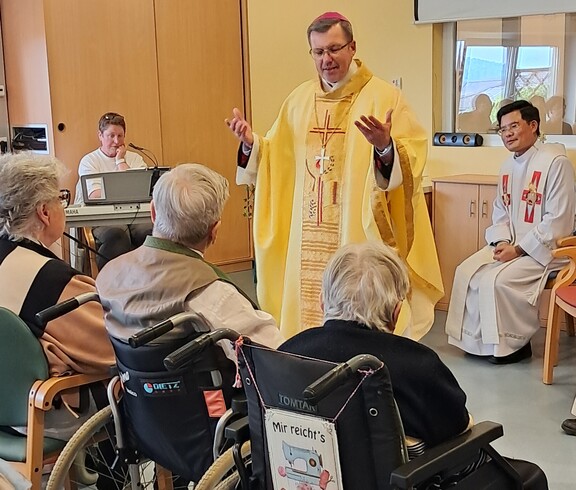 Feier eines Gottesdienstes im Pflegewohnhaus, bei dem der Weihbischof vor den sitzenden Bewohnerinnen und Bewohnern im Gemeinschaftsraum steht.