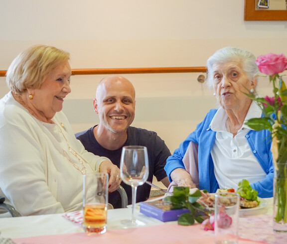 Zwei Damen und einen Mann sitzen gemeinsam an einen Tisch und lächeln. Der Tisch ist mit Blumen und Gläsern wegen der Muttertagsfeier festlich dekoriert,