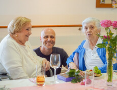 Zwei Damen und einen Mann sitzen gemeinsam an einen Tisch und lächeln. Der Tisch ist mit Blumen und Gläsern wegen der Muttertagsfeier festlich dekoriert,