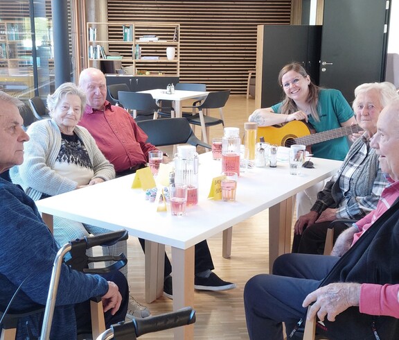 Das Bild zeigt eine gemeinsame Runde im Tageszentrum, in der mehrere ältere Personen an einem Tisch zusammensitzen, Getränke trinken und durch musikalische Begleitung eine betreute Gruppenaktivität erleben.