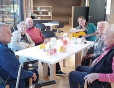 Das Bild zeigt eine gemeinsame Runde im Tageszentrum, in der mehrere ältere Personen an einem Tisch zusammensitzen, Getränke trinken und durch musikalische Begleitung eine betreute Gruppenaktivität erleben.