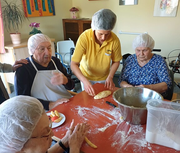 Bewohnerinnen des Pflegewohnhauses formen gemeinsam mit einer Mitarbeiterin frische Germteigfiguren. Am großen Tisch wird geknetet, geformt und gelacht, während Mehl, Teig und Backutensilien für eine lebendige, fröhliche Backrunde sorgen.