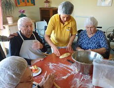 Bewohnerinnen des Pflegewohnhauses formen gemeinsam mit einer Mitarbeiterin frische Germteigfiguren. Am großen Tisch wird geknetet, geformt und gelacht, während Mehl, Teig und Backutensilien für eine lebendige, fröhliche Backrunde sorgen.