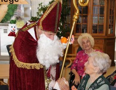 Nikolaus überreicht einer sitzenden Bewohnerin ein kleines Geschenk. Beide lächeln, im Hintergrund ist eine weitere Bewohnerin zu sehen.