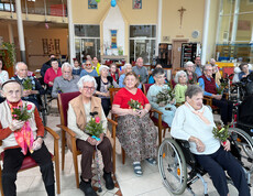 Bewohner*innen sitzen im Gemeinschaftsraum und nehmen an einer österlichen Messe mit Palmzweigen teil.