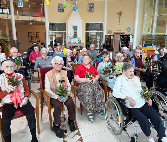 Bewohner*innen sitzen im Gemeinschaftsraum und nehmen an einer österlichen Messe mit Palmzweigen teil.