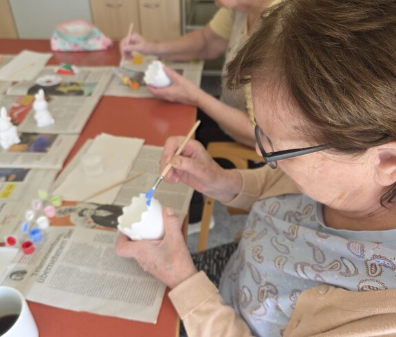 Bewohnerinnen des Pflegewohnhauses bemalen gemeinsam Keramikfiguren und gestalten farbenfrohe Osterdekorationen an einem Arbeitstisch.