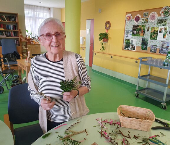 Eine Bewohnerin des Pflegewohnhauses steht am Basteltisch und hält frisch gebundene Palmbuschen in den Händen. Vor ihr liegen Zweige, Bänder und Werkzeuge, während im freundlich gestalteten Aufenthaltsbereich emsig weitergearbeitet wird.