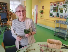 Eine Bewohnerin des Pflegewohnhauses steht am Basteltisch und hält frisch gebundene Palmbuschen in den Händen. Vor ihr liegen Zweige, Bänder und Werkzeuge, während im freundlich gestalteten Aufenthaltsbereich emsig weitergearbeitet wird.