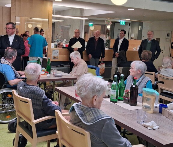 Bewohnerinnen und Bewohner des Caritas Pflegewohnhauses sitzen gemeinsam im Aufenthaltsraum, während der Weihbischof vor der Gruppe spricht; im Hintergrund stehen weitere Gäste und Mitarbeitende im Bereich der Rezeption.