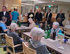 Bewohnerinnen und Bewohner des Caritas Pflegewohnhauses sitzen gemeinsam im Aufenthaltsraum, während der Weihbischof vor der Gruppe spricht; im Hintergrund stehen weitere Gäste und Mitarbeitende im Bereich der Rezeption.