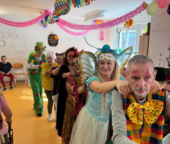 Verkleidete Bewohner*innen machen im geschmückten Raum eine Polonaise unter bunten Girlanden.