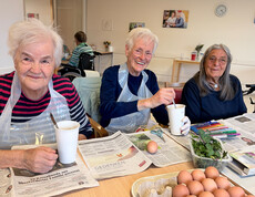 Bewohner*innen färben gemeinsam Ostereier an einem Tisch, Eierkartons, Farben und Zeitungspapier liegen bereit.