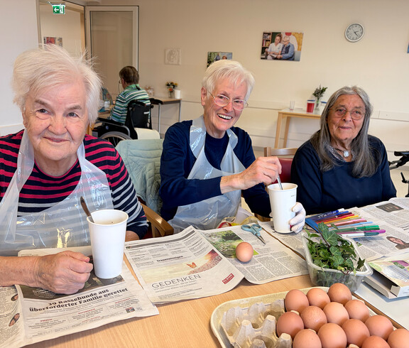 Bewohner*innen färben gemeinsam Ostereier an einem Tisch, Eierkartons, Farben und Zeitungspapier liegen bereit.