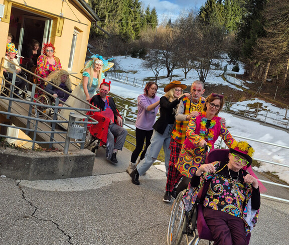 Verkleidete Bewohner*innen kommen in einer Polonaise die Rampe des Hauses hinunter, umgeben von winterlicher Landschaft.