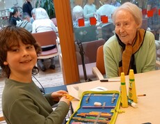 An einem Tisch im Pflegewohnhaus sitzen ein Kind und eine ältere Bewohnerin gemeinsam beim Basteln mit Stiften und Papier, während im Hintergrund weitere Bewohnerinnen und Bewohner sowie Kinder im Gemeinschaftsraum zu sehen sind.