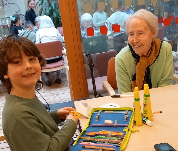 An einem Tisch im Pflegewohnhaus sitzen ein Kind und eine ältere Bewohnerin gemeinsam beim Basteln mit Stiften und Papier, während im Hintergrund weitere Bewohnerinnen und Bewohner sowie Kinder im Gemeinschaftsraum zu sehen sind.