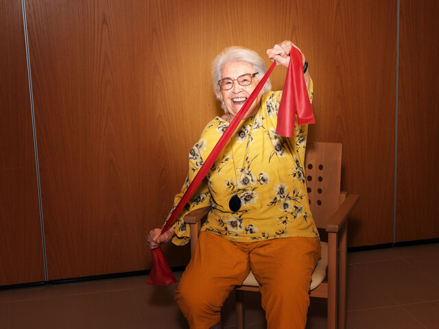 Eine alte Dame zieht ein Theraband in die Länge für eine Übung, sie lächelt in die Kamera.