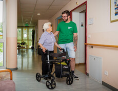 Eine Dame mit Rollator lächelt einem Pfleger zu, während sie im Gang des Pflegewohnhauses stehen.
