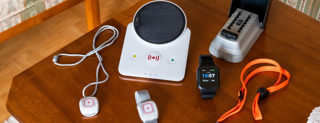 Mehrere Geräte des Caritas-Notrufsystems liegen auf einem Holztisch: eine Basisstation, ein Telefon, kleine Notrufknöpfe mit Verbindungskabel sowie ein mobiler Notrufanhänger mit Trageband.