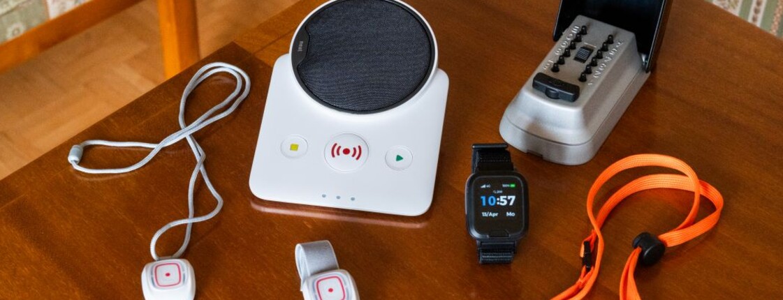Mehrere Geräte des Caritas-Notrufsystems liegen auf einem Holztisch: eine Basisstation, ein Telefon, kleine Notrufknöpfe mit Verbindungskabel sowie ein mobiler Notrufanhänger mit Trageband.