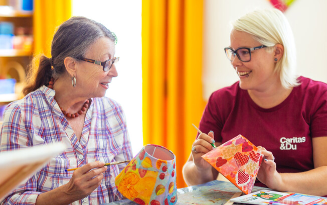 Eine ältere Dame und eine Pflegefachassistentin basteln gemeinsam, bunte Lampenschirme mit der "Serviettentechnik"