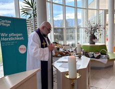 Bewohner*innen sitzen im Gemeinschaftsraum und nehmen an einer österlichen Messe mit Palmzweigen teil.